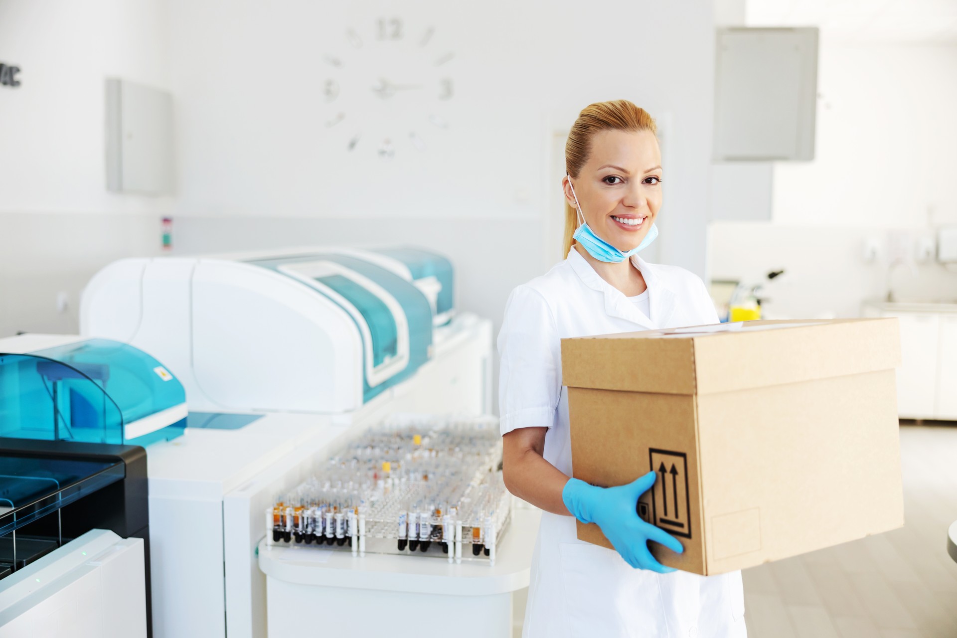 Attractive positive blond lab assistant carrying box with vaccines for Covid 19. Laboratory interior.